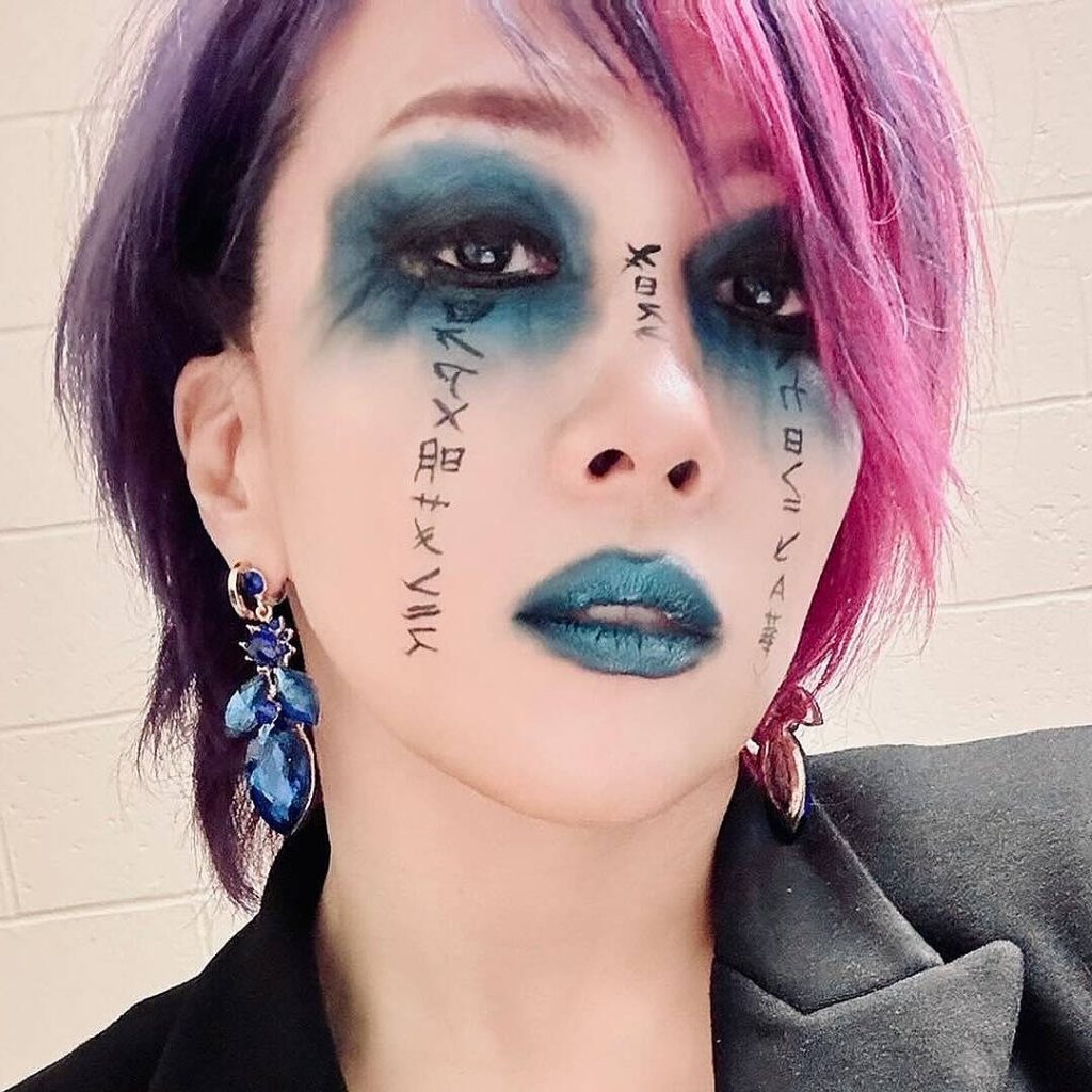 A headshot of professional wrestler Asuka. She is shown from the shoulders up, looking directly at the camera with a confident expression. Her hair is short and dyed purple on the left side and magenta on the right. Her face is painted with blue eyeshadow and lipstick, and she has black, symbol-like markings painted on both cheeks. She is wearing a black blazer and large blue earrings.