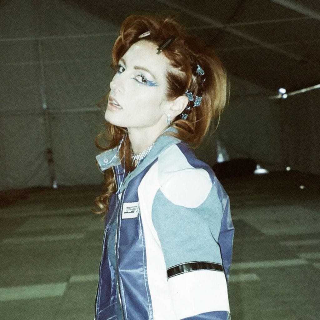 A professional portrait of professional wrestler Becky Lynch. She has long, wavy reddish-blonde hair styled in two pigtails with blue clips, and she is wearing a blue, white, and black racing-style jacket over a sparkly top. She has bold blue eye makeup and is looking back over her shoulder at the camera. The background is a dark, empty warehouse.