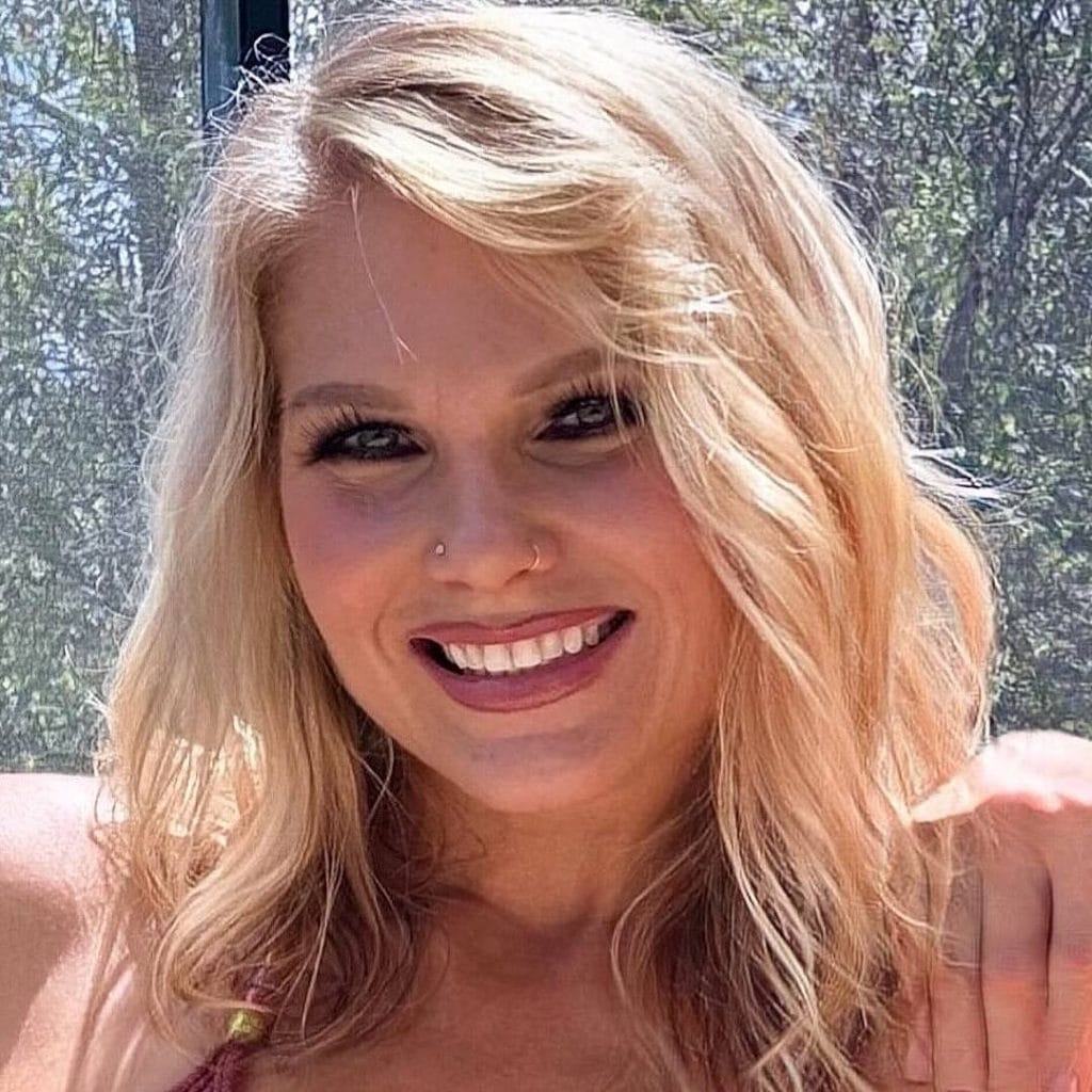 A headshot-style photo of professional wrestler Beth Phoenix. She is a woman with wavy, dirty blonde hair and is smiling broadly at the camera. She is wearing a dark tank top and a small nose stud, and a tree with green leaves is blurred in the background.