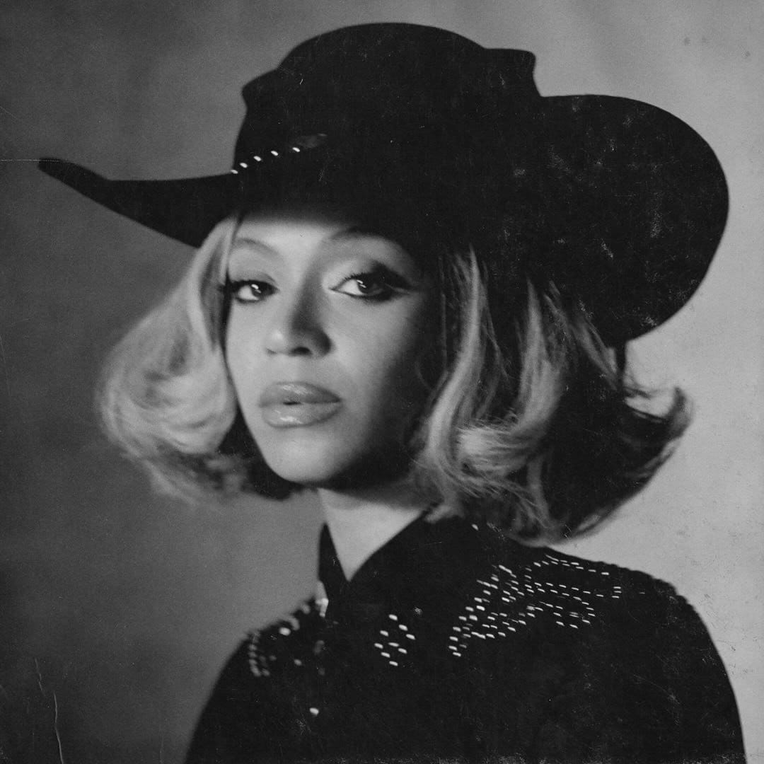 A black and white portrait of Beyoncé. She is wearing a wide-brimmed cowboy hat and a dark jacket with beaded detailing. She has a wavy bob hairstyle and a serious expression. The photo has a grainy, vintage quality.