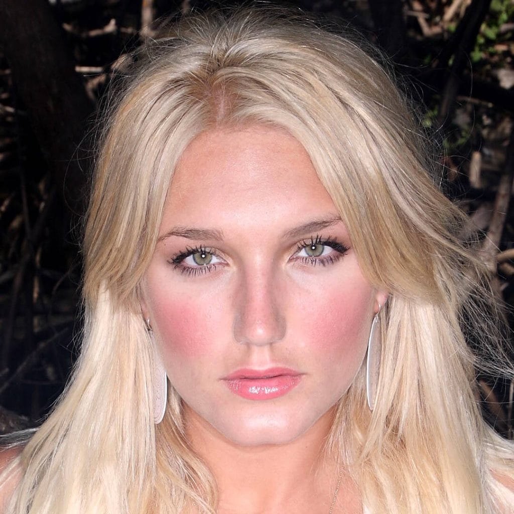 A close-up portrait of Brooke Hogan looking directly at the camera. She has long, blonde hair, bright green eyes, and is wearing large, silver, oval-shaped earrings.