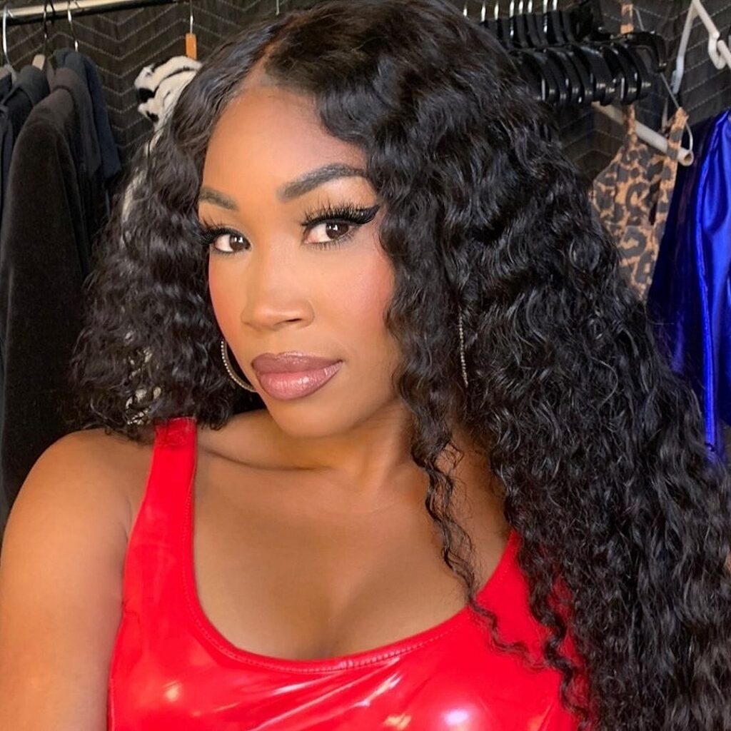 Close-up selfie of professional wrestler and reality star Cameron (Ariane Andrew) with long, wet-look curly hair, wearing a shiny red latex or PVC tank top with a dark, cluttered background.
