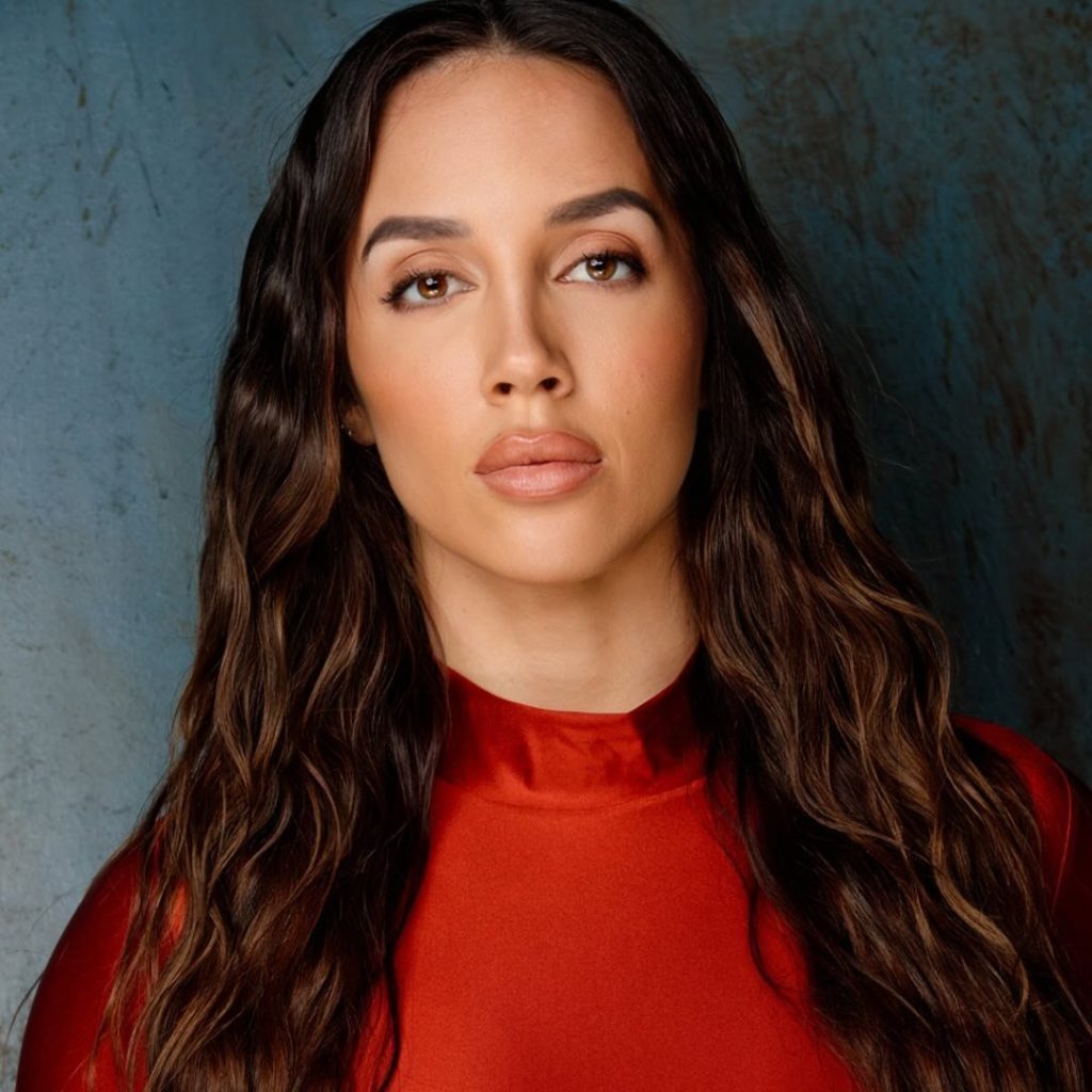 Headshot of professional wrestler Chelsea Green with long, dark, wavy hair and intense gaze, wearing a red high-neck top against a mottled blue and gray background.