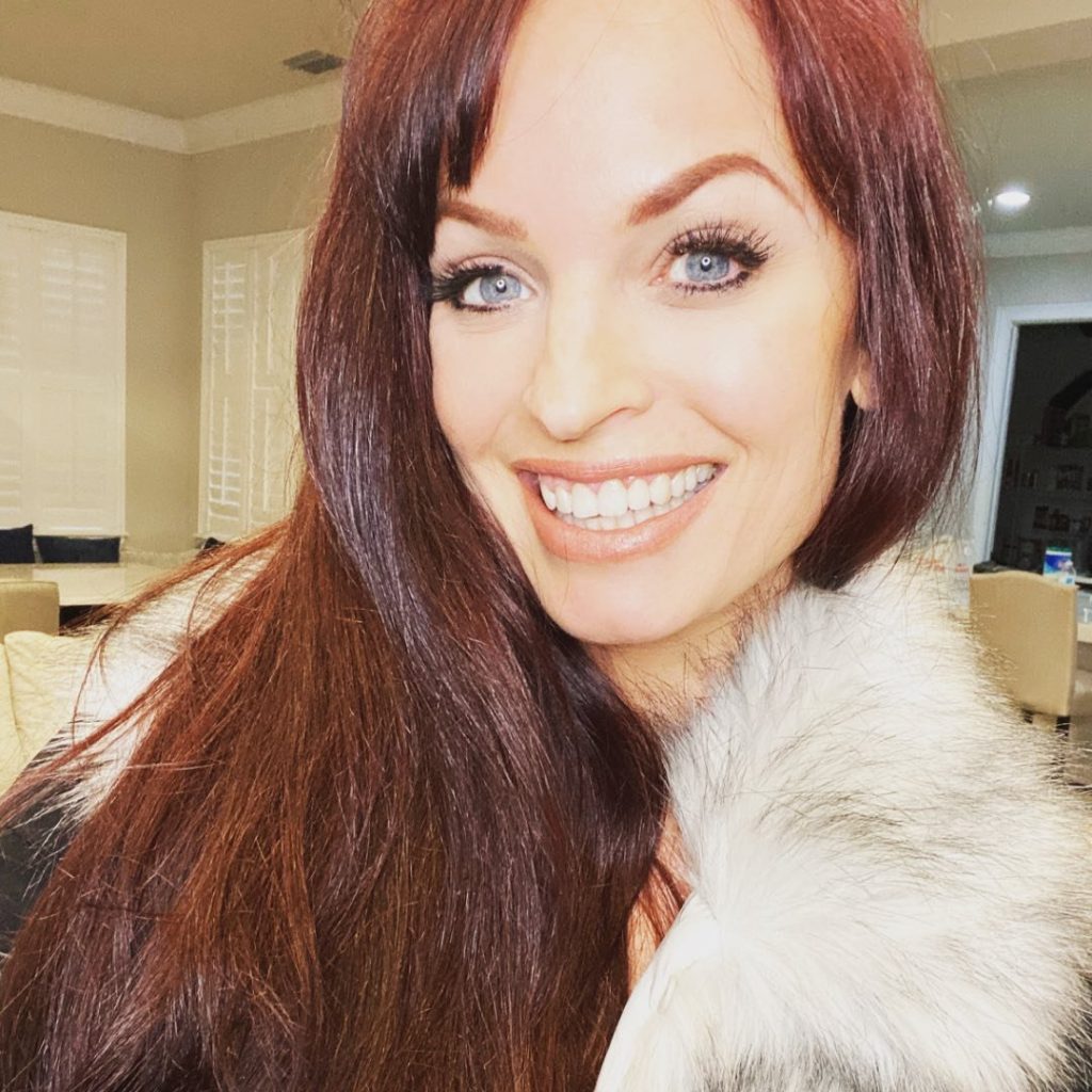 Close-up smiling selfie of Christy Hemme with long auburn hair, bright blue eyes, and wearing a white faux fur shawl or coat indoors with a blurred living area in the background.