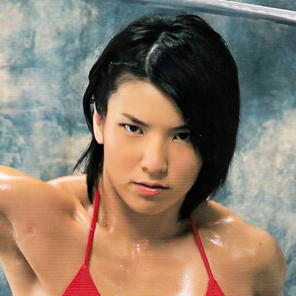 Close-up portrait of professional wrestler Hikaru Shida with short dark hair, wearing a red bikini top, looking intently at the camera, with sweat visible on her shoulders and a blurred blue background.