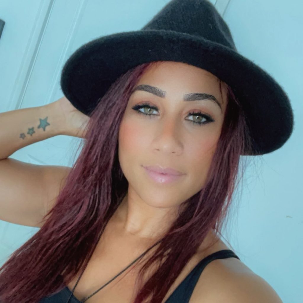 Selfie of reality TV personality Hoopz (Nicole Alexander) wearing a black fedora or wide-brimmed hat and a black tank top, with reddish-brown hair and a three-star tattoo on her left arm.