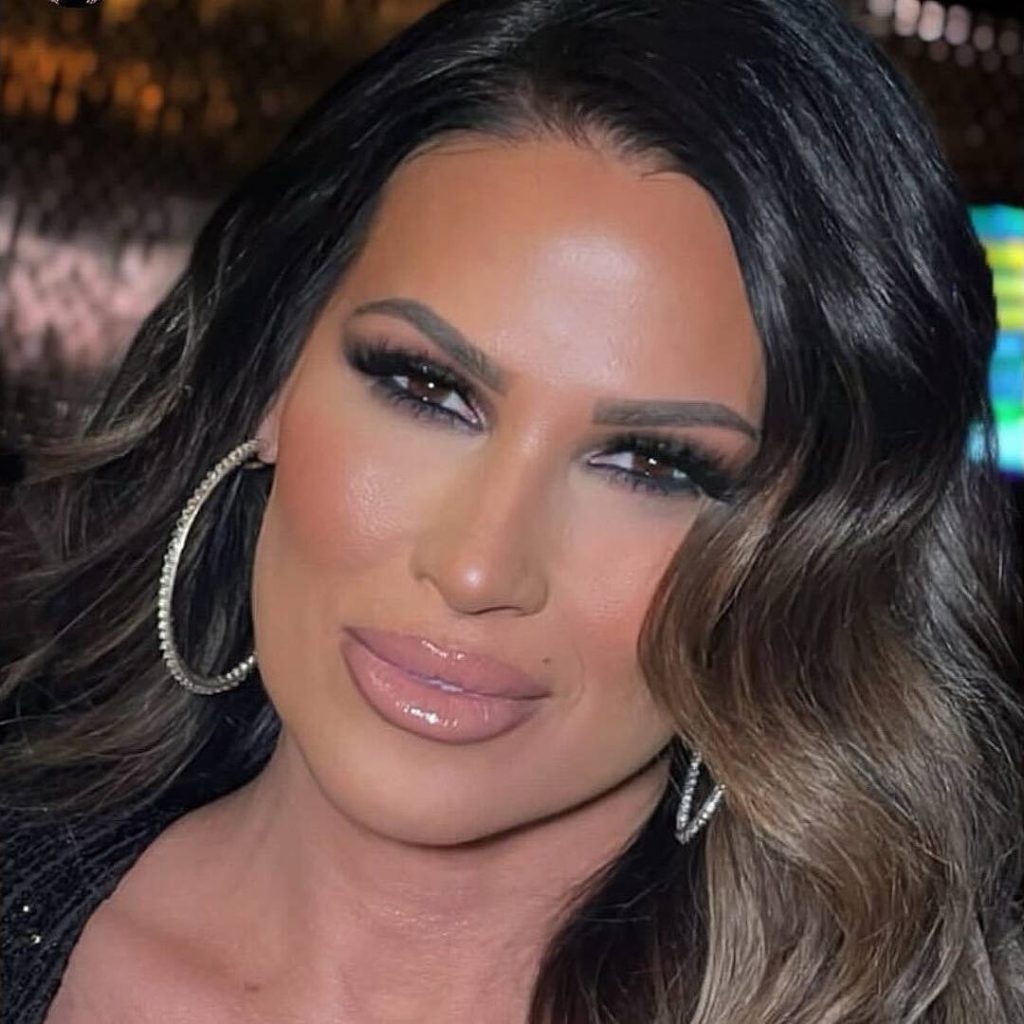 Close-up portrait of wrestling personality Karen Jarrett with long, dark, wavy hair, wearing a black top and large diamond hoop earrings, featuring heavy eye makeup and glossy nude lips.