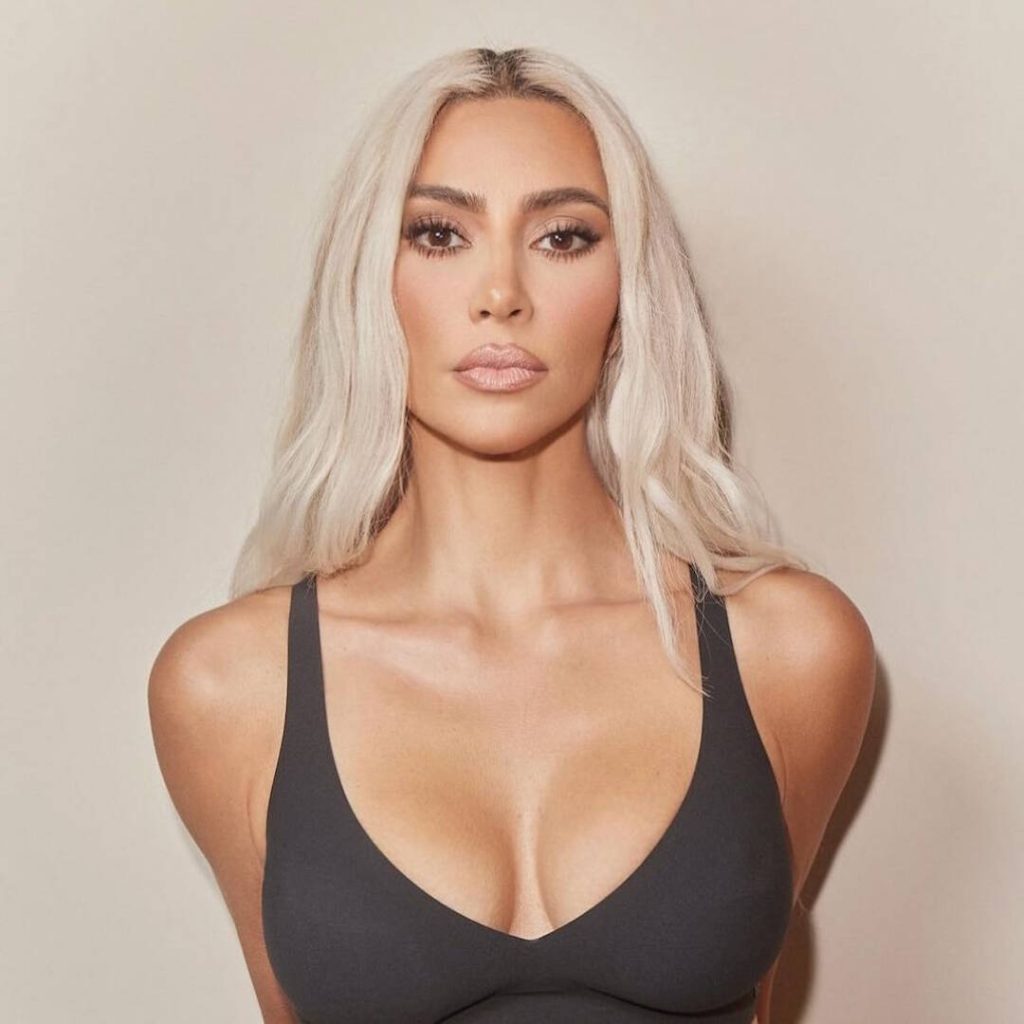 Close-up portrait of reality TV star and entrepreneur Kim Kardashian with long, wavy platinum blonde hair, wearing a black low-cut tank top and staring directly at the camera.