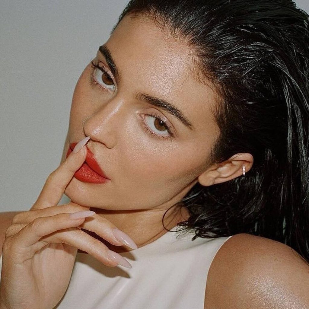 Close-up portrait of model and entrepreneur Kylie Jenner with wet, slicked-back dark hair, red matte lipstick, and a nude manicure, touching her lips with her finger.