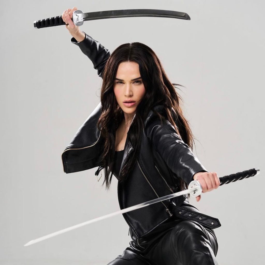 Professional wrestler Lana (C.J. Perry) poses aggressively in a black leather jacket and pants, holding two silver katanas in a defensive stance on a gray background.