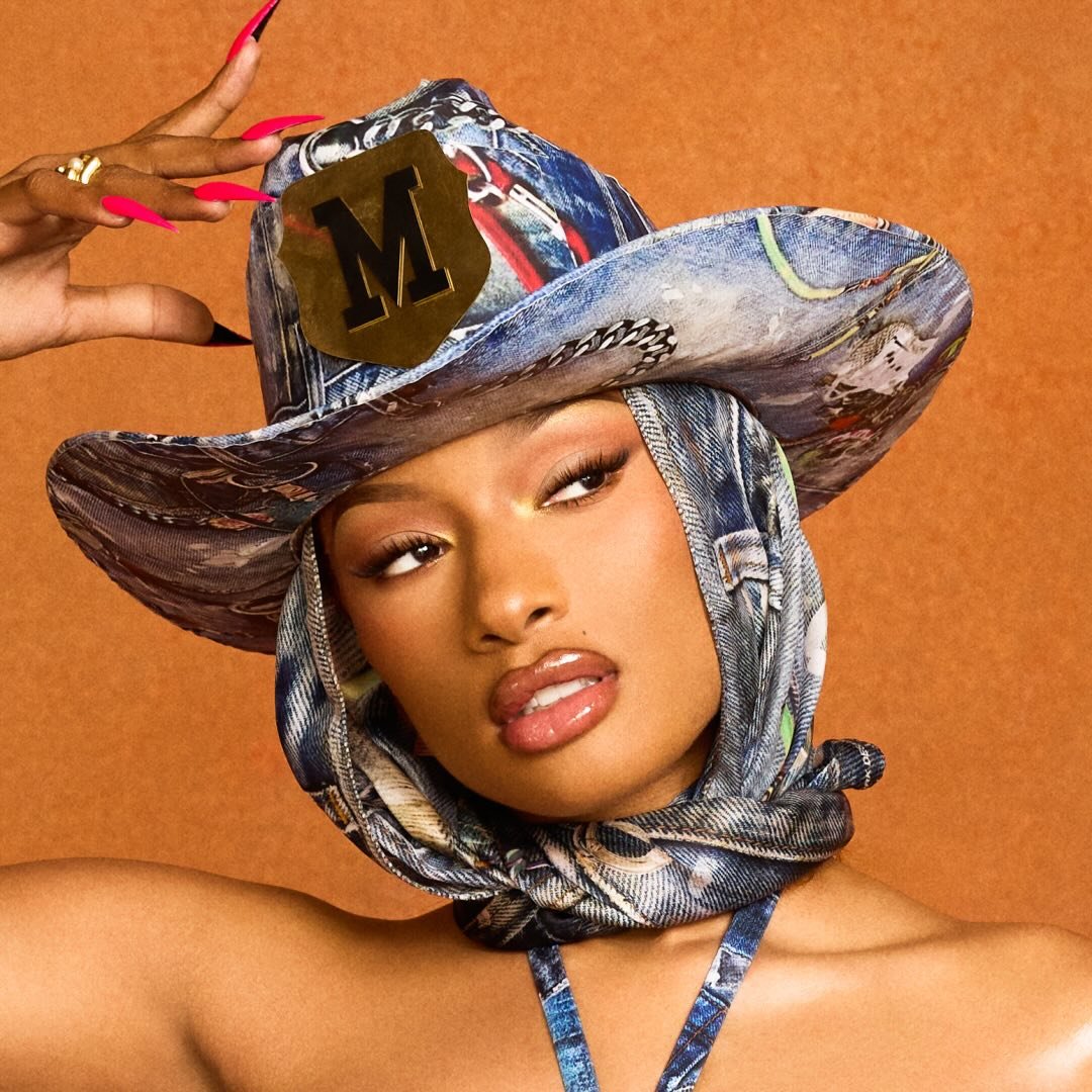 Close-up portrait of rapper Megan Thee Stallion wearing a denim-patterned cowboy hat with a scarf tied under her chin, featuring bright pink manicure and a gold "M" emblem on the hat.