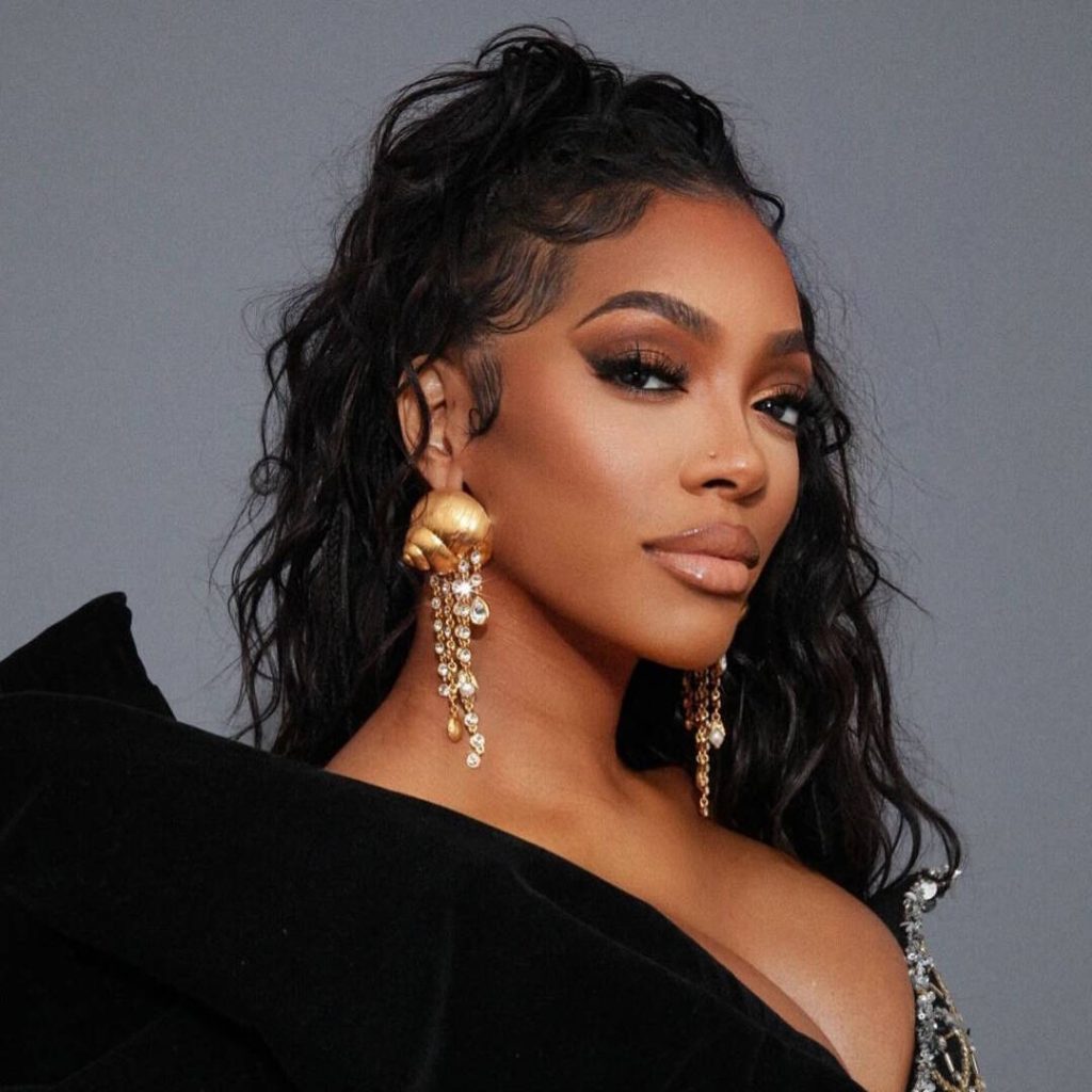 Close-up portrait of reality TV personality Porsha Williams with long, dark, wavy hair pulled into a high half-ponytail, wearing a black velvet gown with a dramatic off-the-shoulder bow, and large gold dangling earrings.