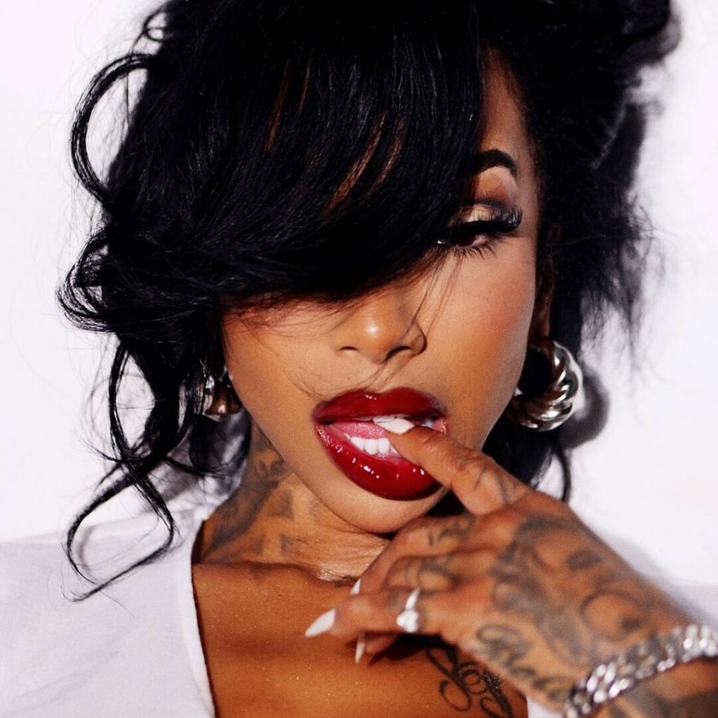 Close-up portrait of the artist Skyy Days with a hand near her glossy red lips, which are biting her finger, featuring dark hair, a white manicure, and visible tattoos on her neck and chest.