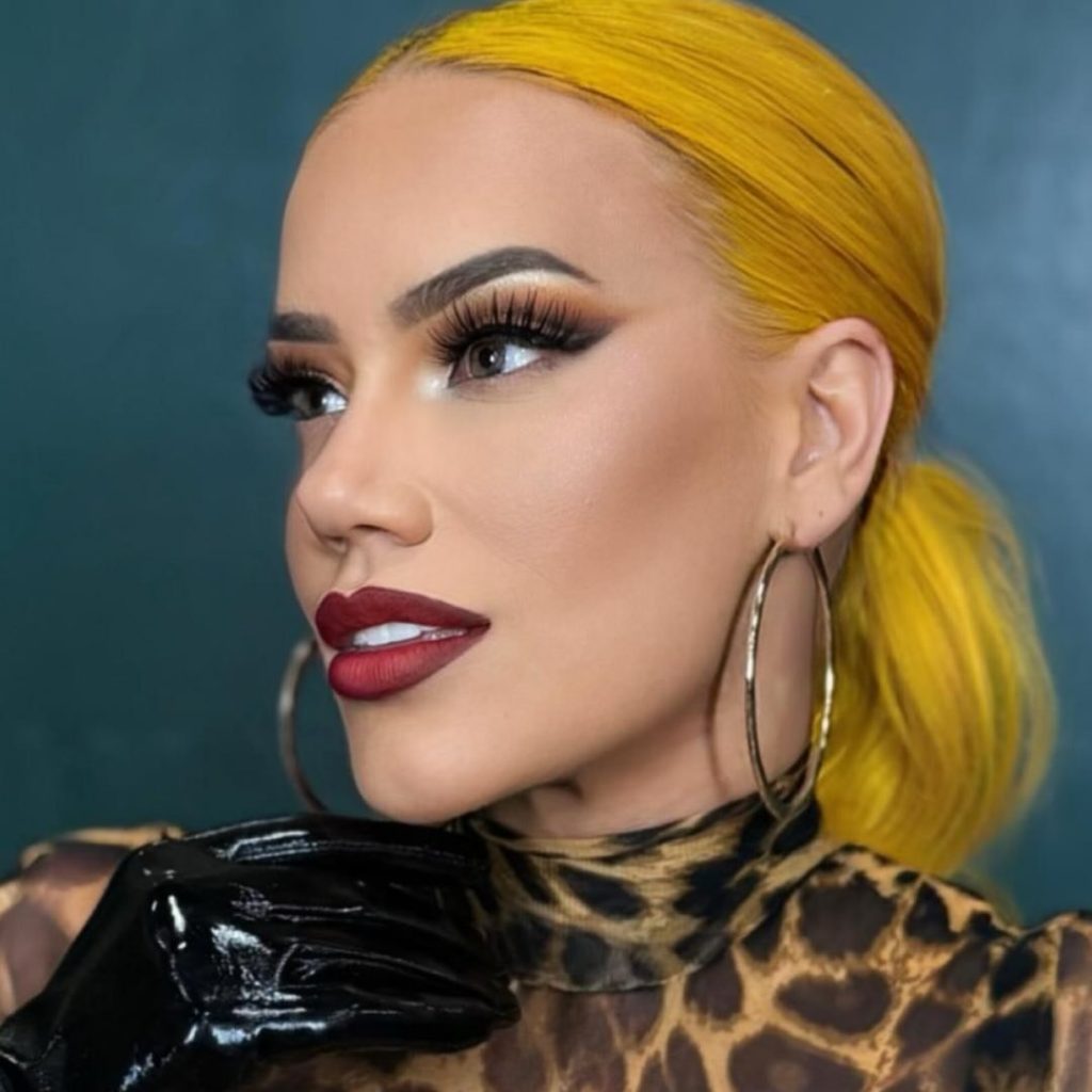 Close-up profile of professional wrestler Taya Valkyrie with bright yellow hair pulled back, wearing a leopard print top and a black patent leather glove, featuring dramatic makeup and large hoop earrings.