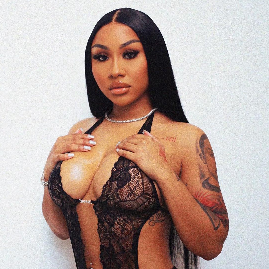 Influencer Ari Fletcher posing in a black lace lingerie bodysuit, featuring a diamond tennis necklace and showcasing her arm tattoos against a plain white background.
