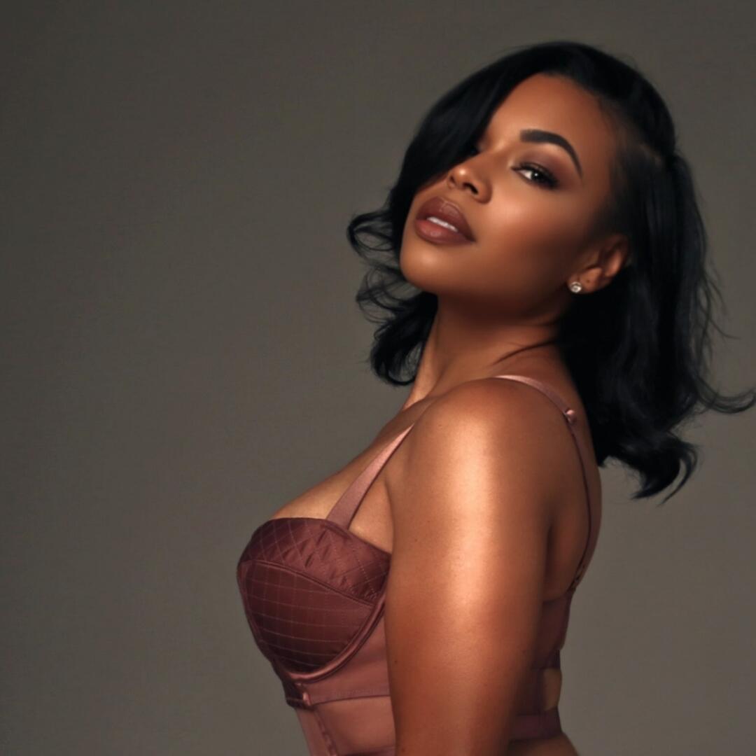 Actress Alexis Fields posing in a side-profile studio portrait, featuring short black wavy hair, a mauve bustier top, and stud earrings against a dark gray background.