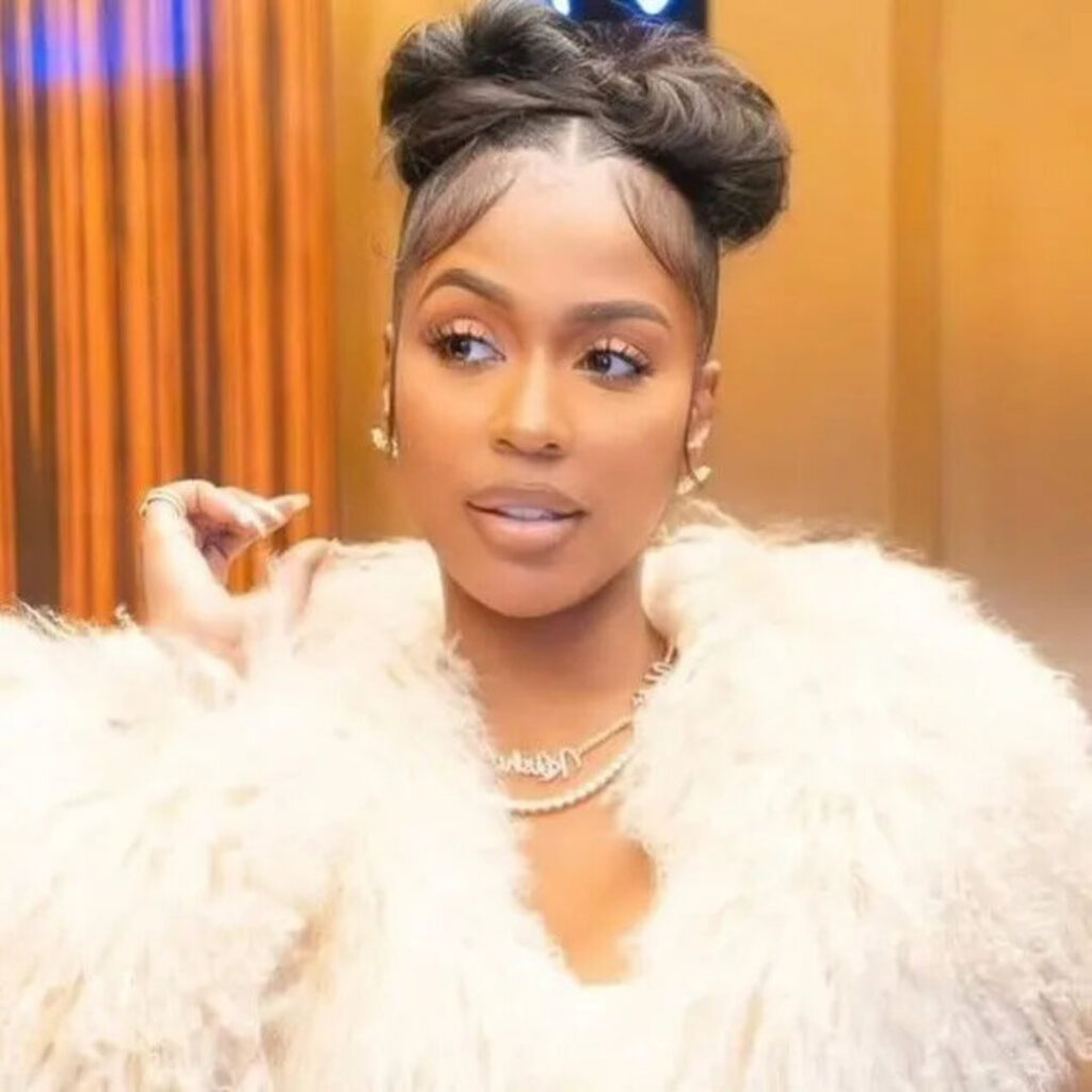 Rapper Kash Doll posing in a close-up portrait, featuring a high braided double bun updo with styled baby hairs, a plush white faux-fur coat, and gold chain necklaces.