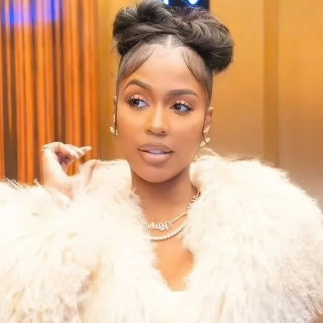 Rapper Kash Doll posing in a close-up portrait, featuring a high braided double bun updo with styled baby hairs, a plush white faux-fur coat, and gold chain necklaces.