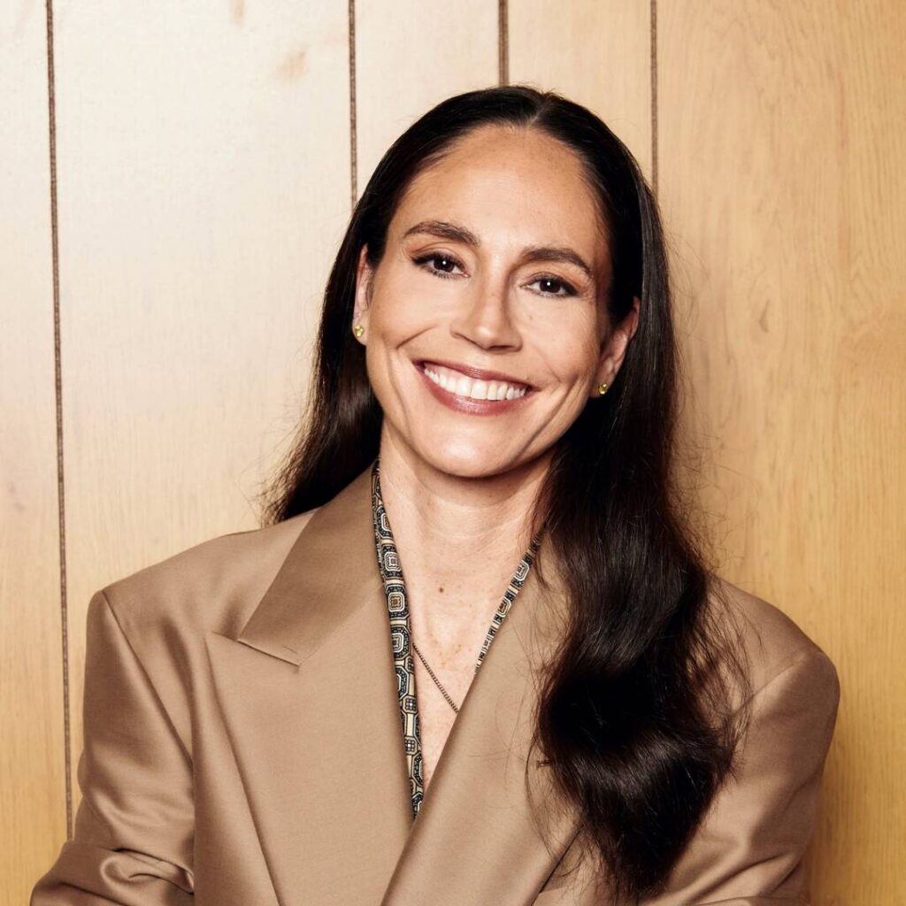 Basketball star Sue Bird posing in a warm studio portrait with dark long hair, a broad smile, a beige blazer, and small stud earrings against a wood-paneled wall.